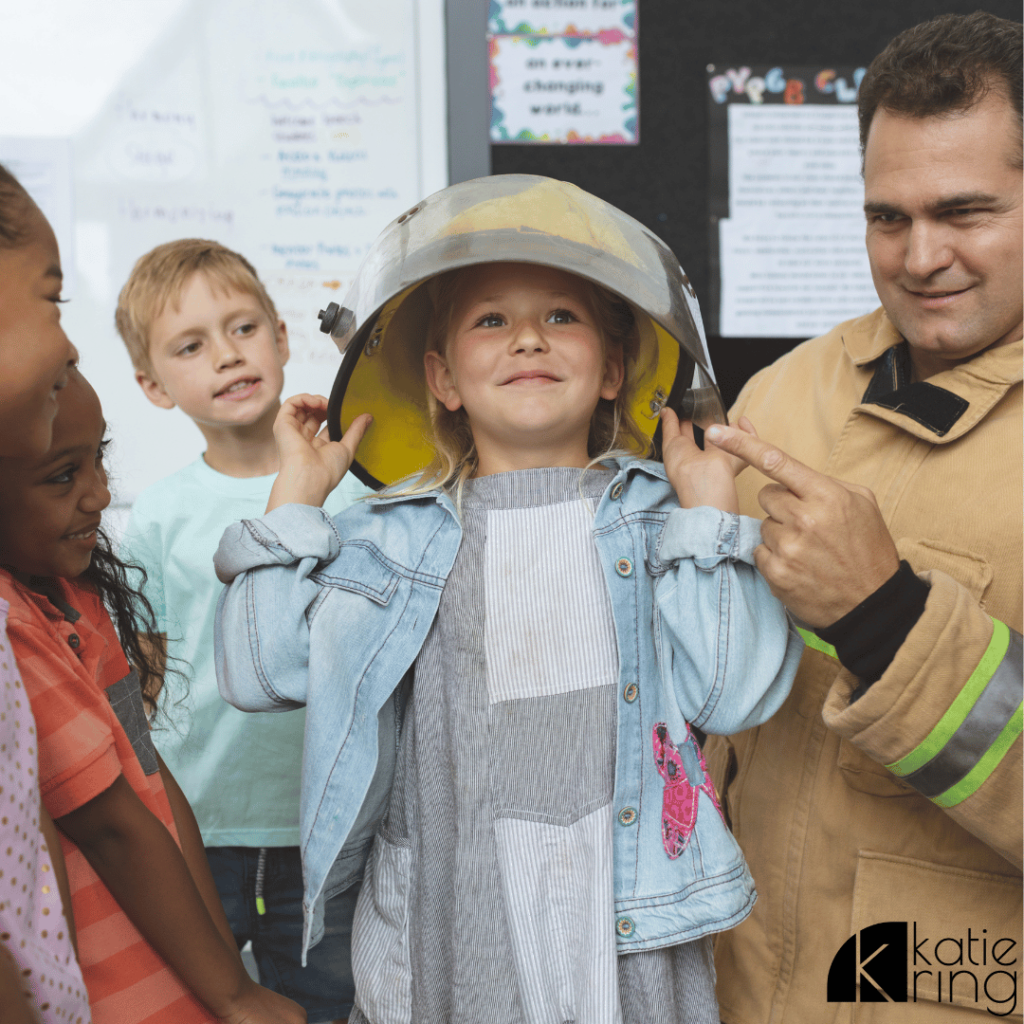 This photo shows a group of elementary age children visiting a fire department. The firemen in the photo is helping a young girl try on the firefighter helmet.
